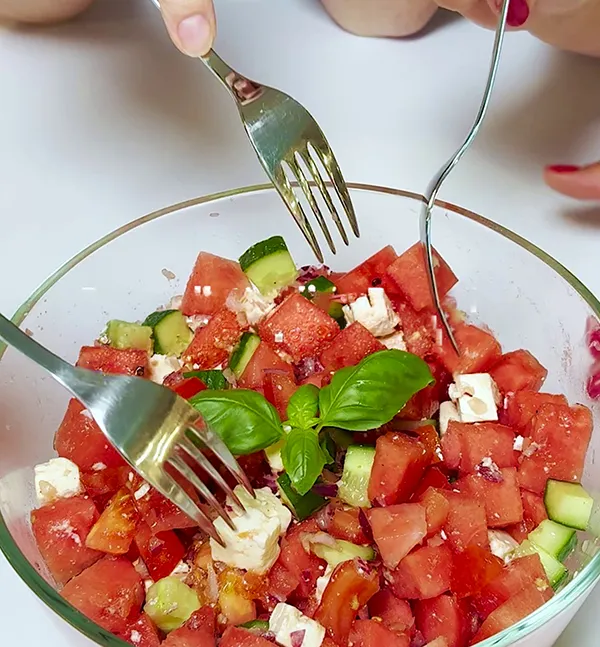 Zwei Gabeln mischen frischen Wassermelonen-Gurken-Salat mit Feta-Käse und Basilikum in einer Glasschüssel