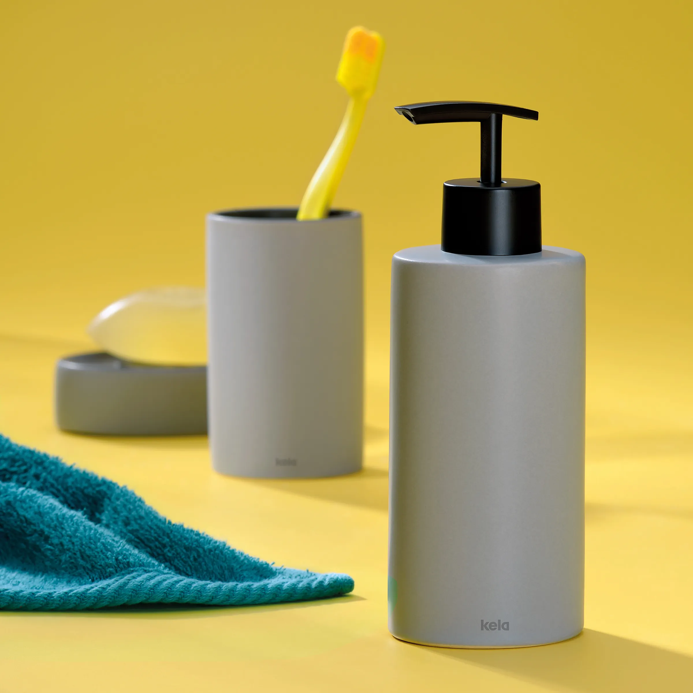 Badezimmer Accessoires Set - Seifenspender und Zahnputzbecher grau Grauer Seifenspender und Zahnputzbecher mit gelber Zahnbürste auf türkisem Handtuch vor gelbem Hintergrund