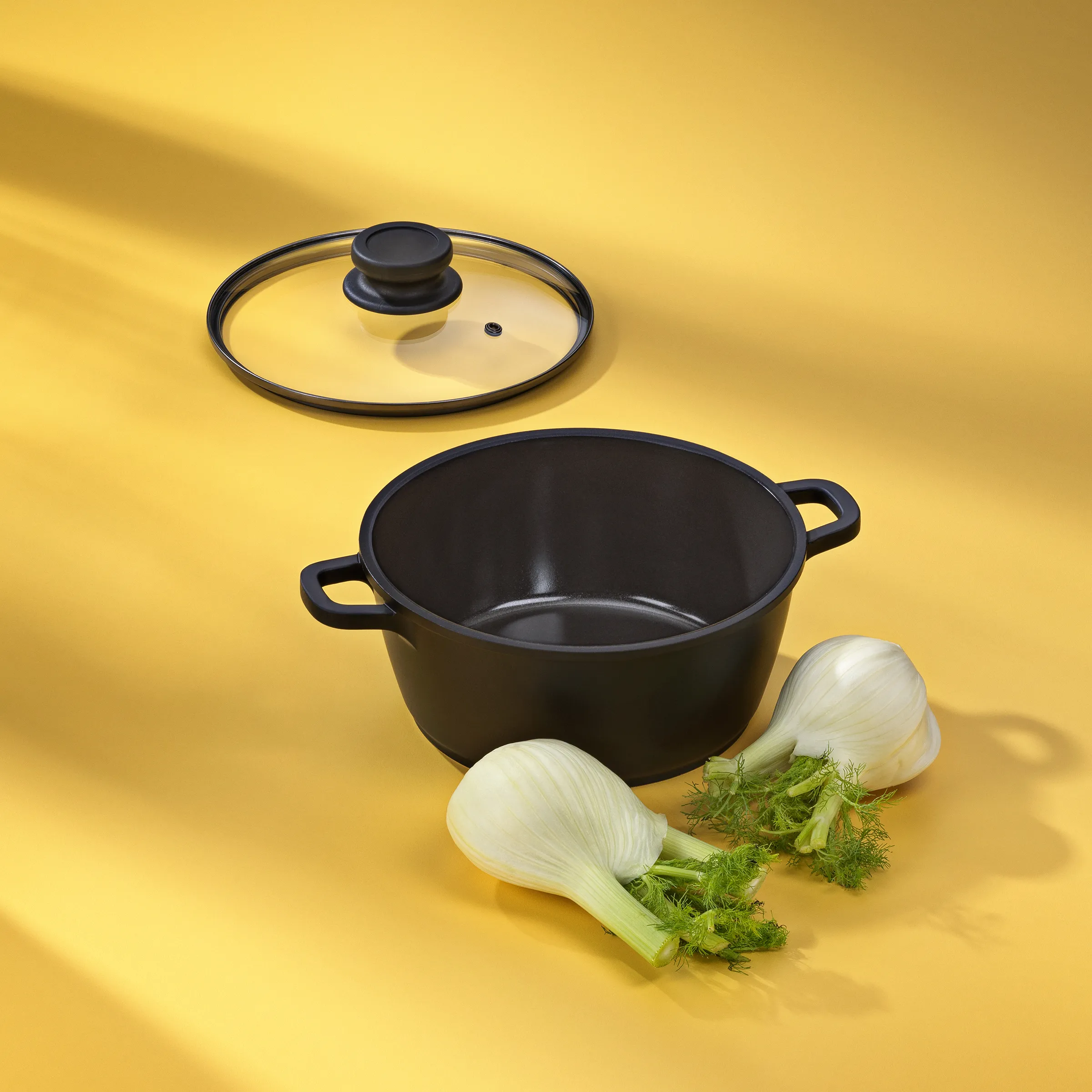 Schwarzer Kochtopf mit Glasdeckel und frischem Fenchel auf gelbem Hintergrund