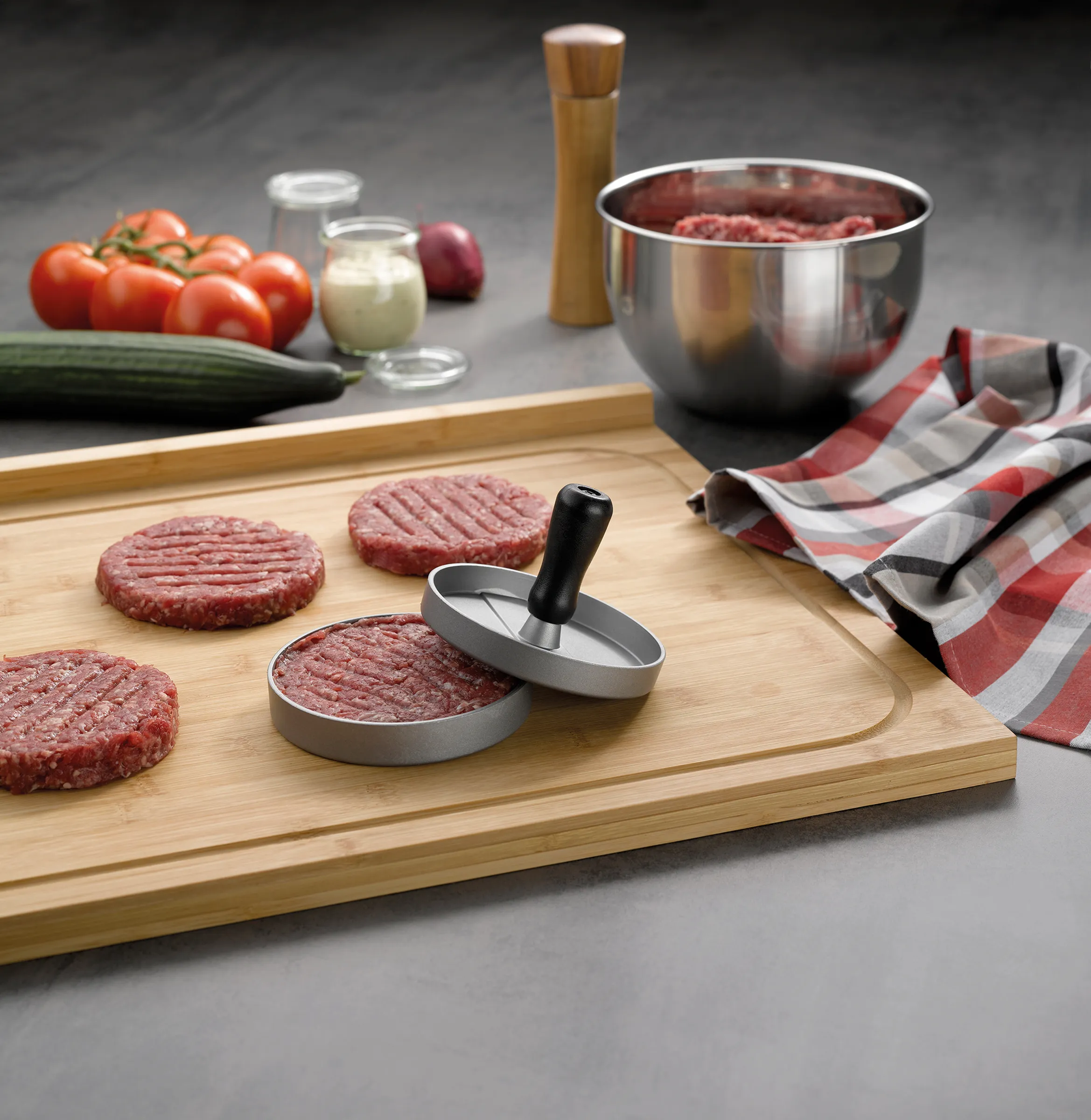 Burgerpresse für perfekte Hamburger-Patties - Küchenhelfer aus Edelstahl Burgerpresse mit rohem Hackfleisch auf Holzbrett, daneben Burger-Patties und Zutaten wie Tomaten und Zucchini