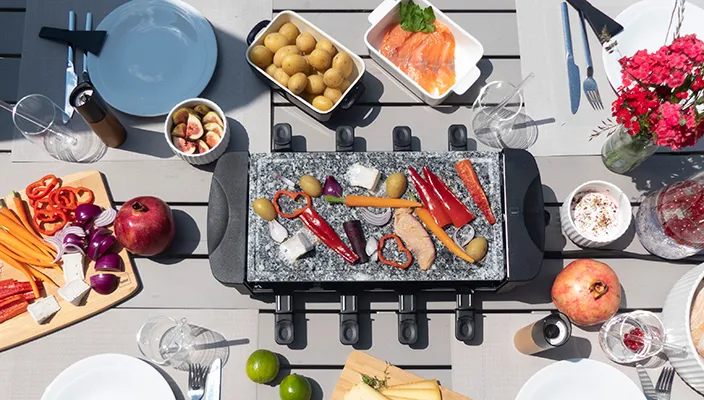Overhead-Ansicht eines Raclette-Grills mit gegrilltem Gemüse und Fleisch, umgeben von frischen Zutaten und Getränken auf weißem Tisch