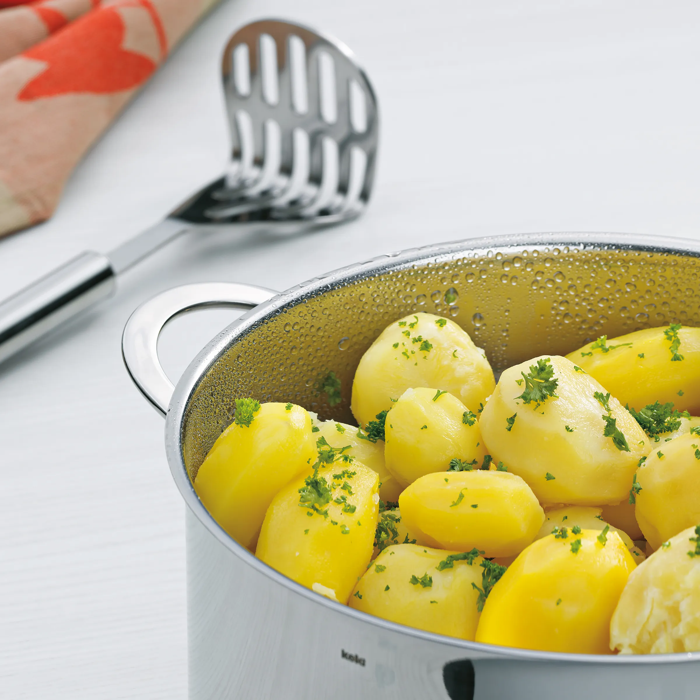 Gekochte Pellkartoffeln mit frischer Petersilie in einem Topf mit Siebeinsatz, daneben Küchenwerkzeuge