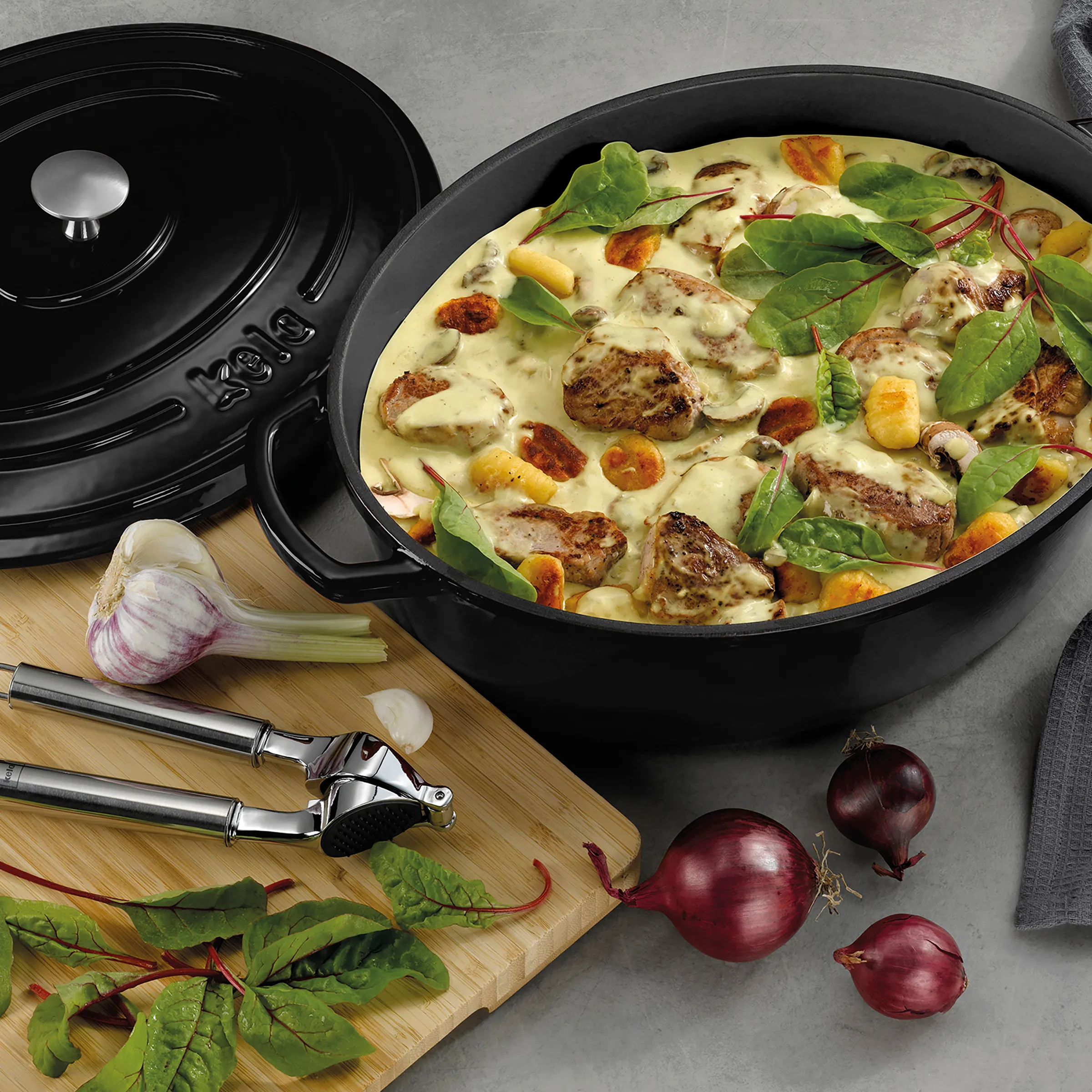 Schmortopf Bräter Rezept: Fleisch Kartoffel Ragout Schwarz glänzender Bräter mit Deckel gefüllt mit Fleischragout, Kartoffeln und Sahnesoße, umgeben von Zwiebeln und frischen Kräutern auf Holzbrett.