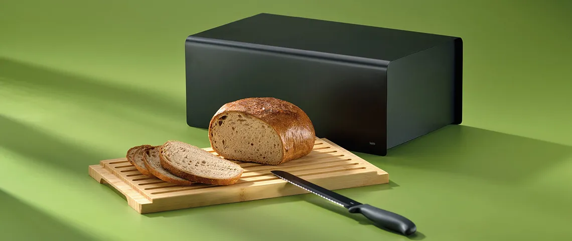 Brotbox schwarz - Brotkasten für frisches Brot Schwarze Brotbox neben Schneidebrett mit Vollkornbrot, Scheiben und Messer auf grünem Hintergrund