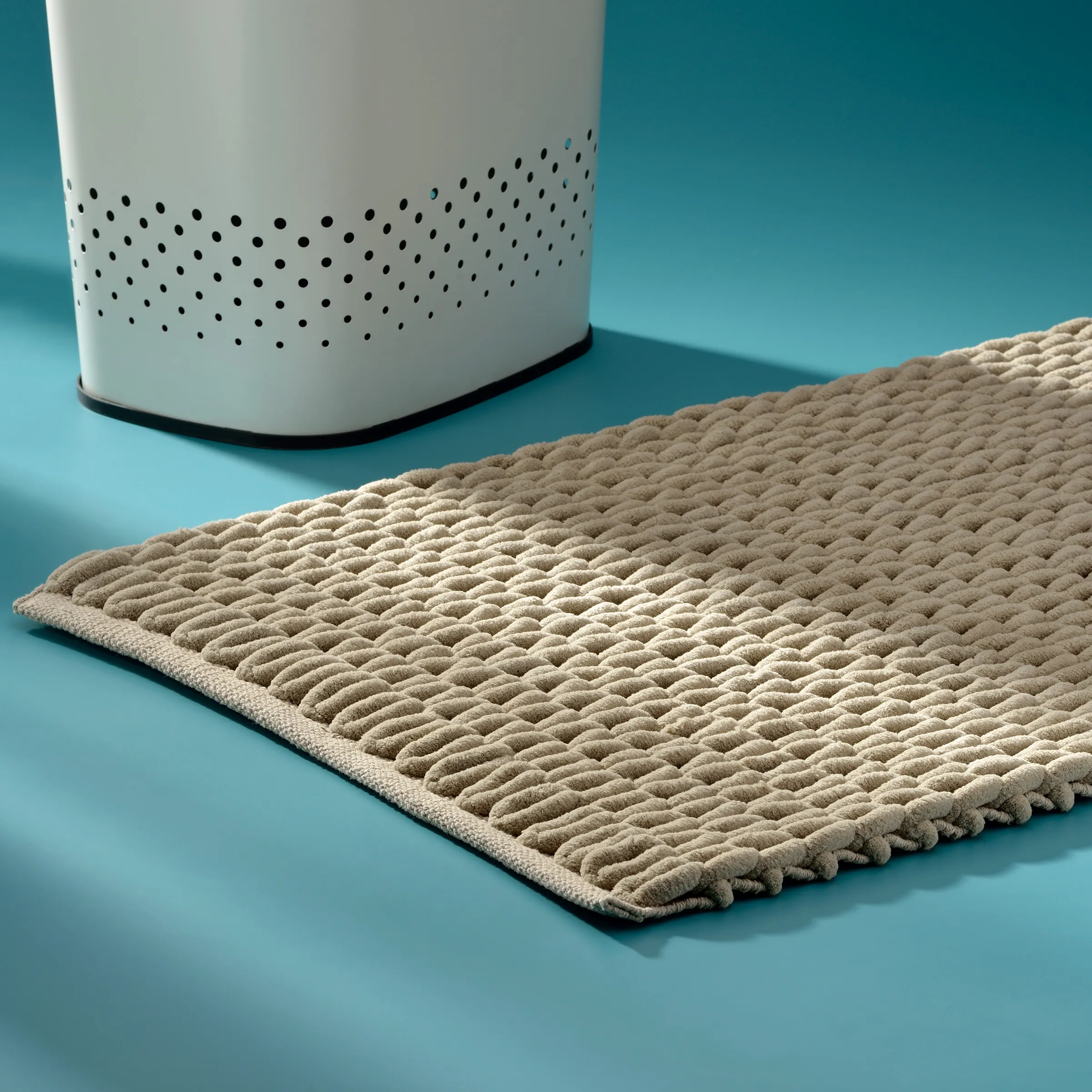 Wäschekorb und Badteppich Set - moderne Badezimmerausstattung Weiße Wäschetrommel mit perforiertem Muster neben beigem strukturiertem Badteppich auf blauem Hintergrund