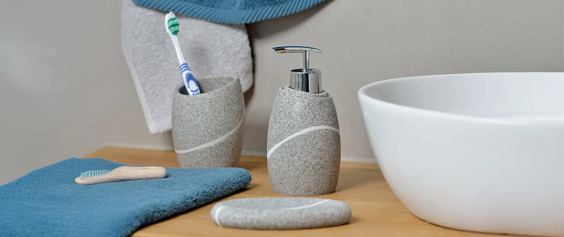 Modernes Badezimmer-Set mit grauer Keramik-Seifenspender, Zahnbürstenhalter, weißer Schüssel und blauen Handtüchern auf Holztisch