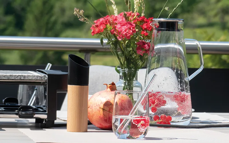 Sommerliche Tischdeko mit Blumen und frischen Früchten Sommerliche Tischszene mit roten Nelken in Vase, Glas mit Erdbeeren, Granatapfel und Trinkflaschen auf Balkon