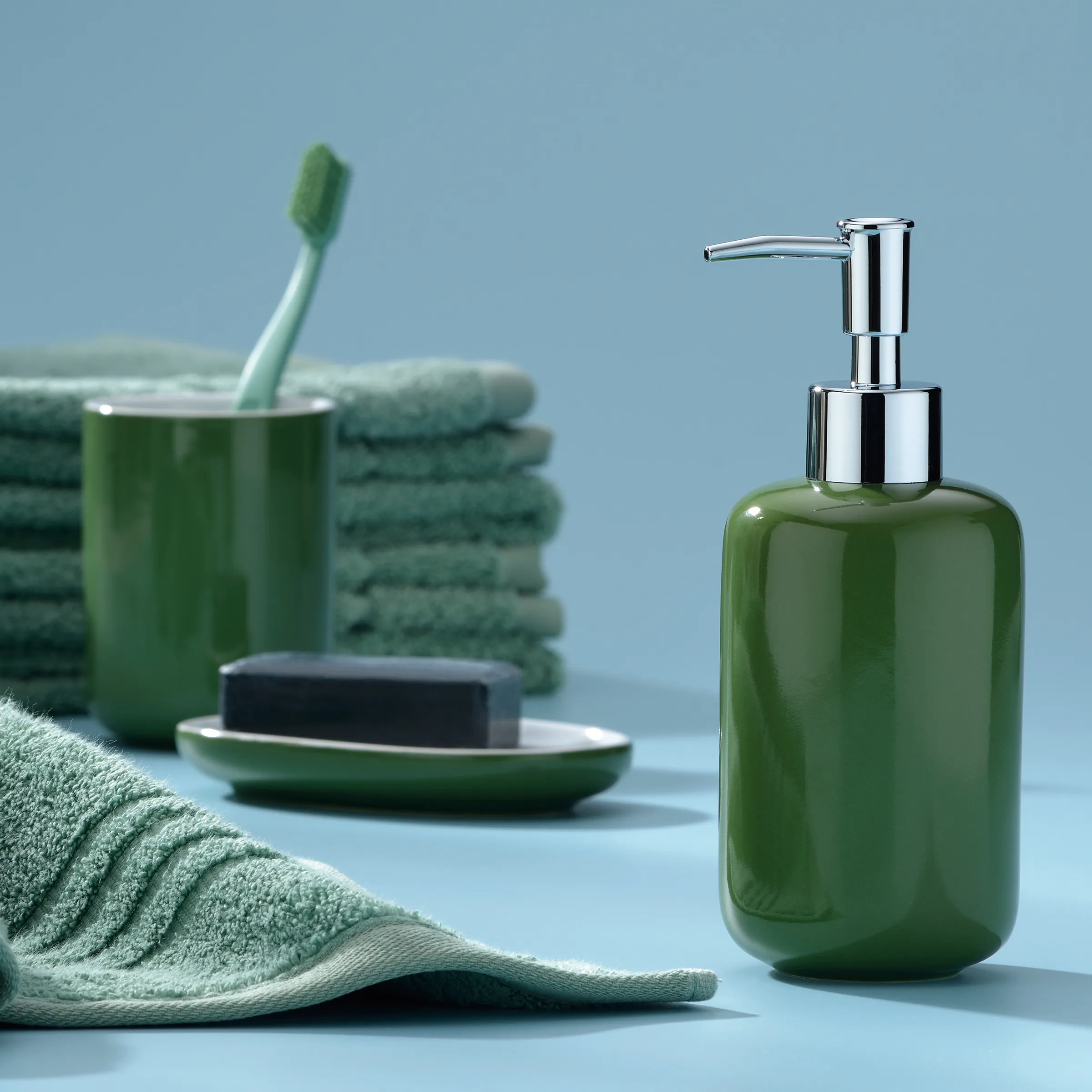 Grünes Badezimmer-Zubehör Set mit Seifenspender und Handtüchern Grüner Seifenspender mit Chrompumpe neben Handtuch, Zahnbürste und Seife auf blauem Hintergrund