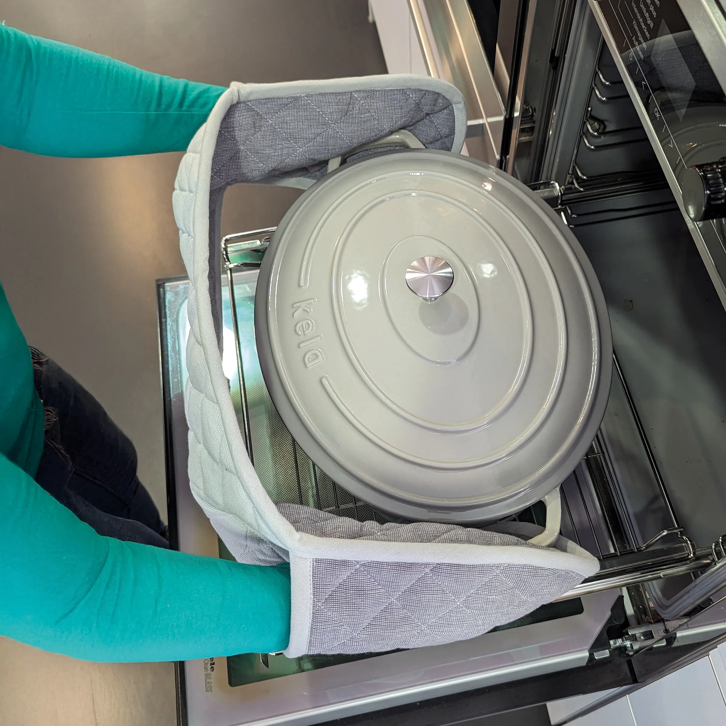 Person mit Topfhandschuhen hält Feld-Kochtopf mit Deckel über geöffnetem Backofen
