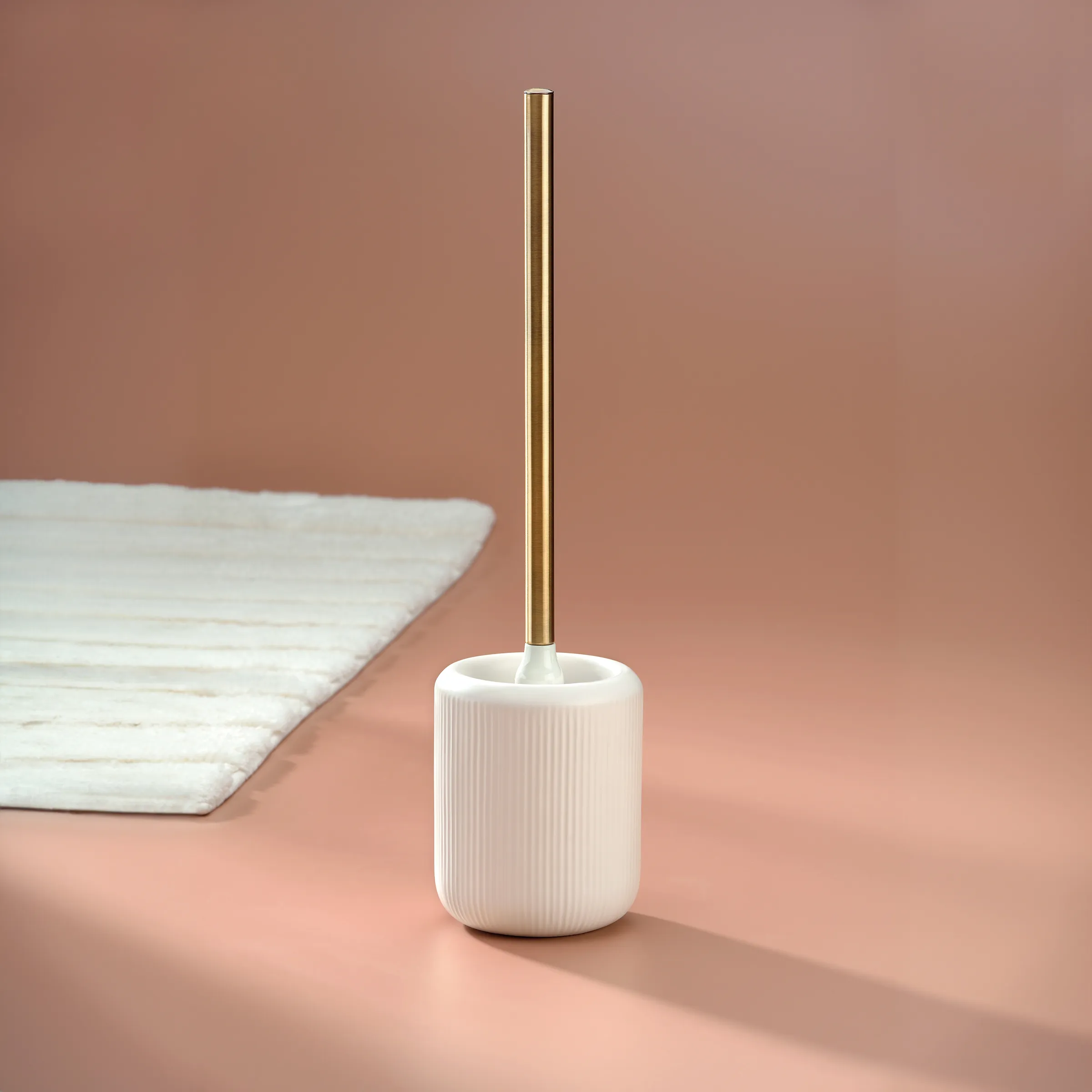 WC-Bürste mit Keramikhalter und Goldgriff - Designer Toilettenbürste Weiße WC-Bürste mit goldenem Griff neben weißen Handtüchern vor rosafarbenem Hintergrund