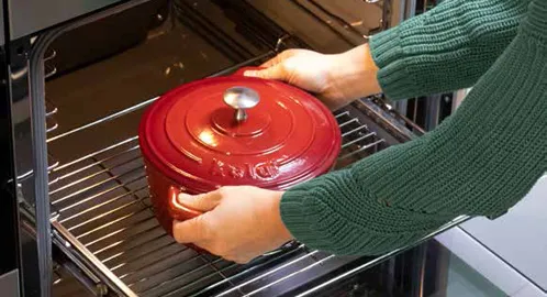 Roter Gusseisen-Bräter wird in Backofen gestellt - Kochgeschirr für Dutch Oven Rezepte Person platziert roten Gusseisen-Bräter mit Deckel in geöffneten Backofen