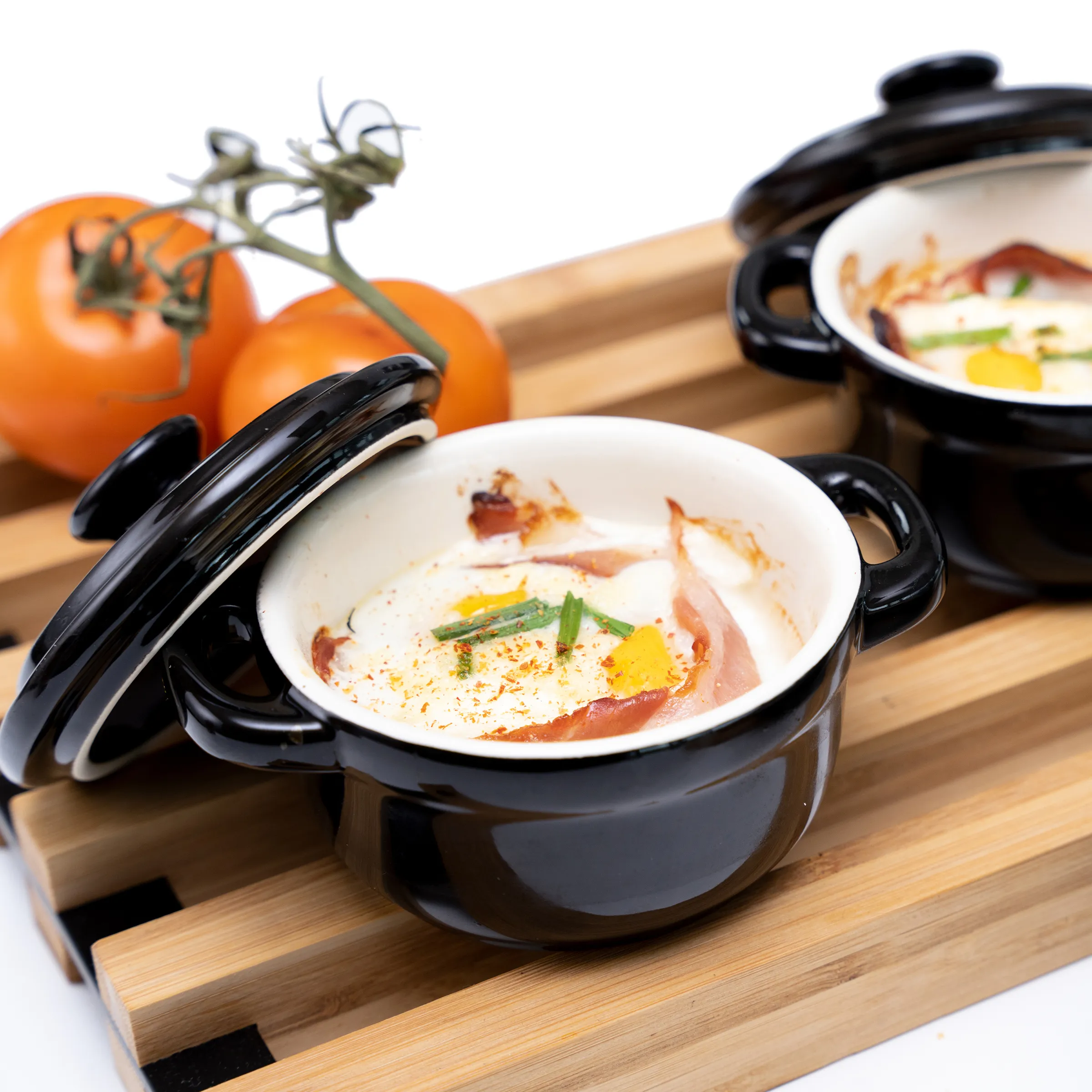Cremige Tomatensuppe in eleganten Keramik-Suppentöpfen Zwei schwarze Keramik-Suppentöpfe mit Deckel auf Holzbrett, gefüllt mit cremiger Suppe, Prosciutto und Gemüse, frische Tomaten im Hintergrund