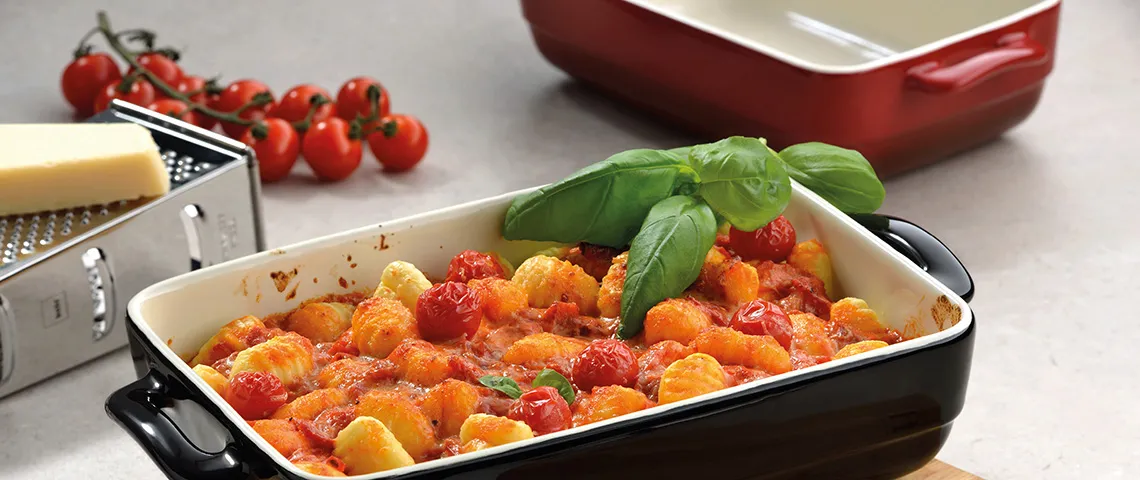 Auflaufform mit gebackenem Gemüse: Tomaten, Kartoffeln und Basilikum, daneben frische Tomaten und Küchenreibe