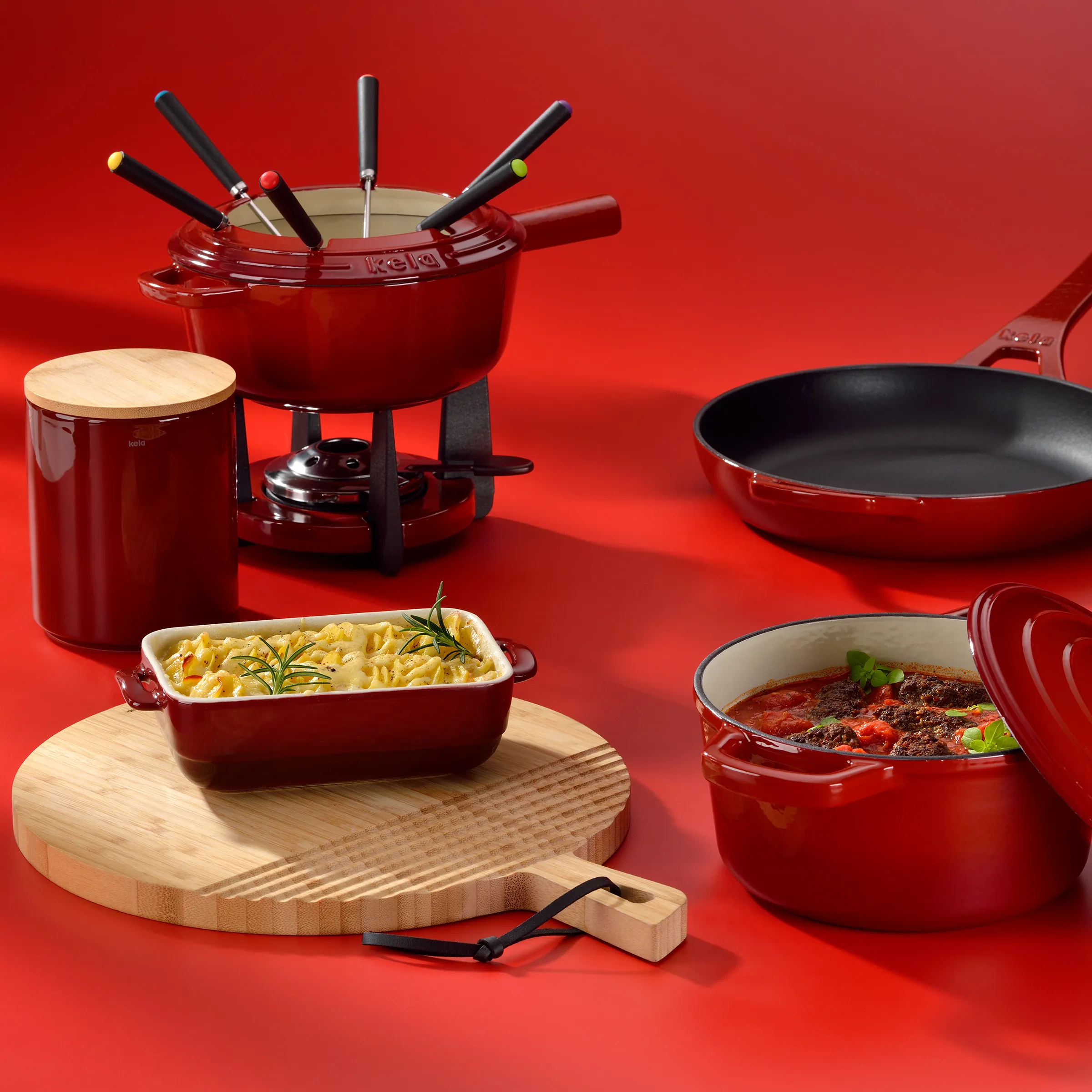Gusseisen Kochgeschirr Set rot - Töpfe, Pfanne und Fondue Rote Kochgeschirr-Set bestehend aus Gusseisen-Töpfen, Pfanne, Fondue-Set und Holzutensilien auf rotem Hintergrund