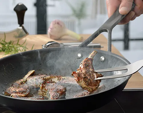 Fleisch braten in Pfanne mit Küchenzange - Steaks perfekt zubereiten Hand hebt gebratenes Fleischstück mit Küchenzange aus schwarzer Pfanne, weitere Steaks brutzeln im Hintergrund