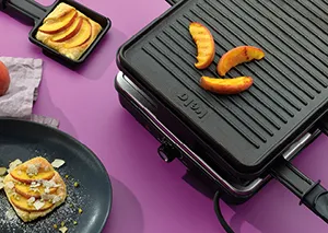 Kontaktgrill mit gegrillten Kartoffeln und Gemüse auf violettem Hintergrund, daneben eine Pfanne mit gegrilltem Essen