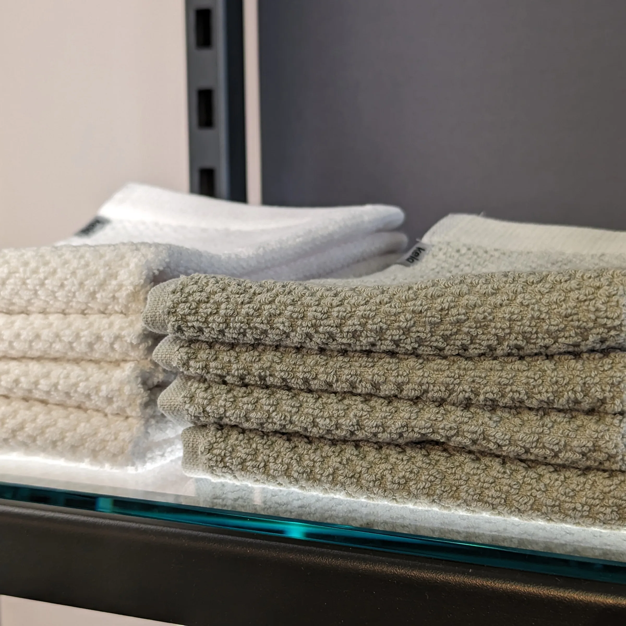 Hochwertige Handtücher Set - Beige und Weiß Gestapelte Handtücher in Beige und Weiß auf einem Glassboard, im Hintergrund ein grauer Behälter.