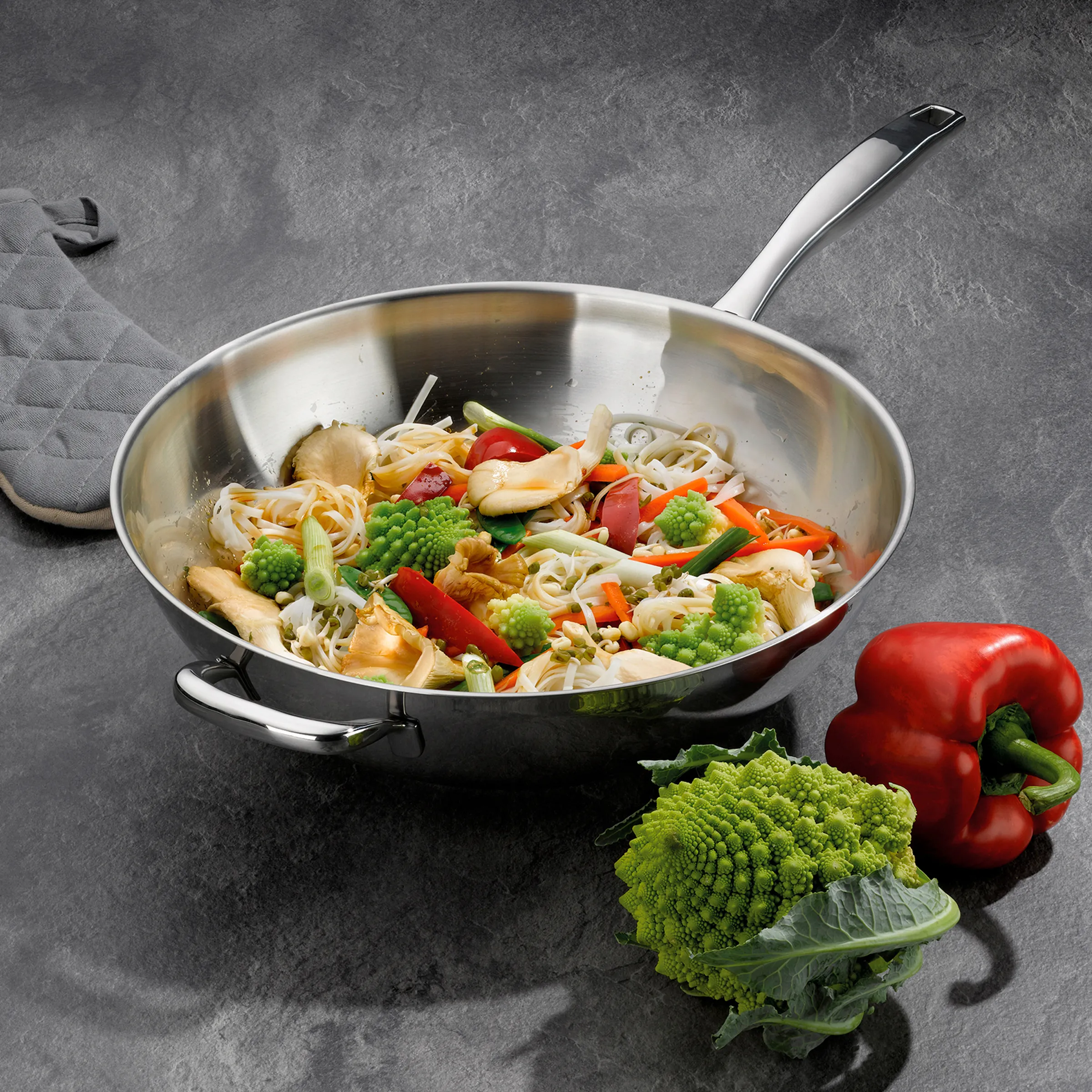 Edelstahl-Wok mit buntem Gemüse-Nudel-Pfannengericht, Paprika und Romanesco auf grauer Arbeitsfläche