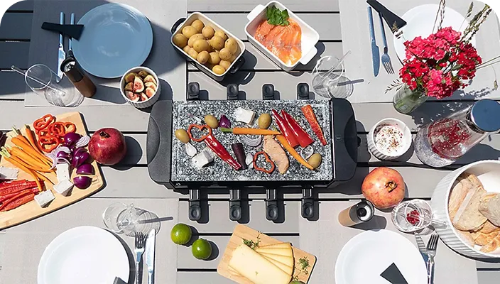 Draufsicht auf ein Raclette-Grill mit Gemüse, Fleisch und Käse auf Serviertellern sowie Besteck und Getränken auf weißem Tisch.