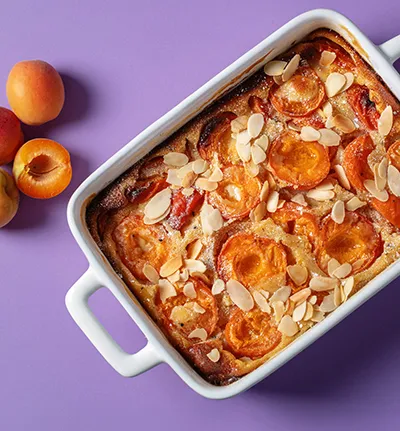 Aprikosen-Mandel-Kuchen in weißer Auflaufform auf violettem Hintergrund mit frischen Aprikosen