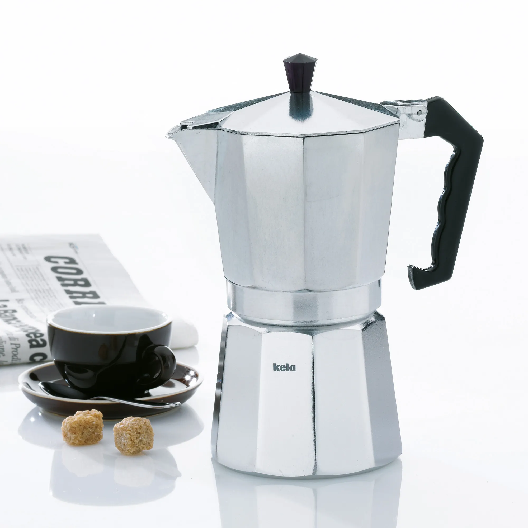 Kela Espressokocher Aluminium mit Espressotasse - Italienische Kaffeezubereitung Kela Espressokocher aus Aluminium mit schwarzem Griff, neben Espressotasse und Zeitung auf weißem Hintergrund
