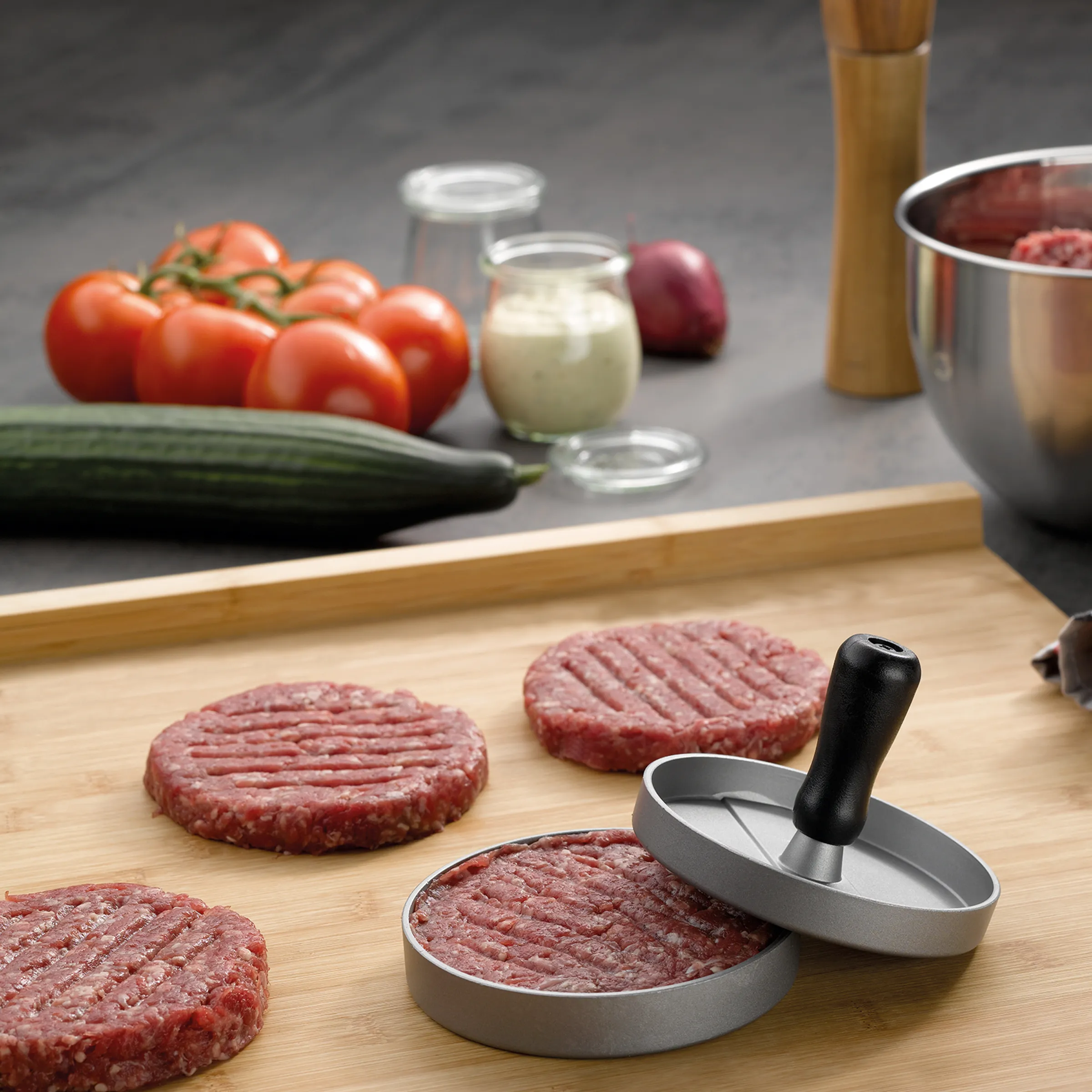 Burger-Presse für perfekte Fleischpatties Burger-Presse mit rohem Fleischpatty auf Holzbrett, daneben weitere Patties und Zutaten wie Tomaten und Zwiebeln