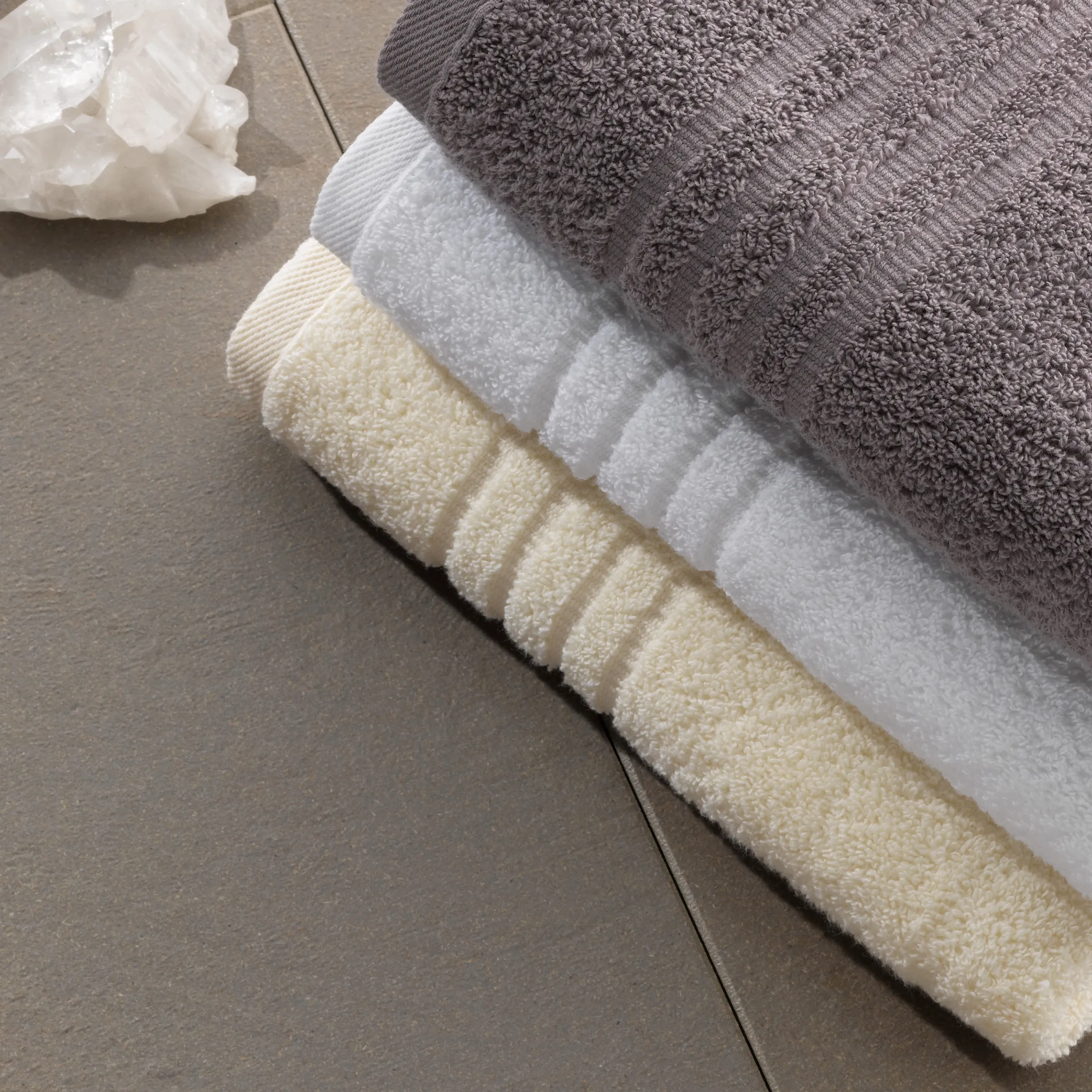 Frotteehandtücher Set in Beige Weiß Grau - Premium Badtextilien Gefaltete Frotteehandtücher in Beige, Weiß und Grau auf hellem Holzboden mit weißer Blumendekoration