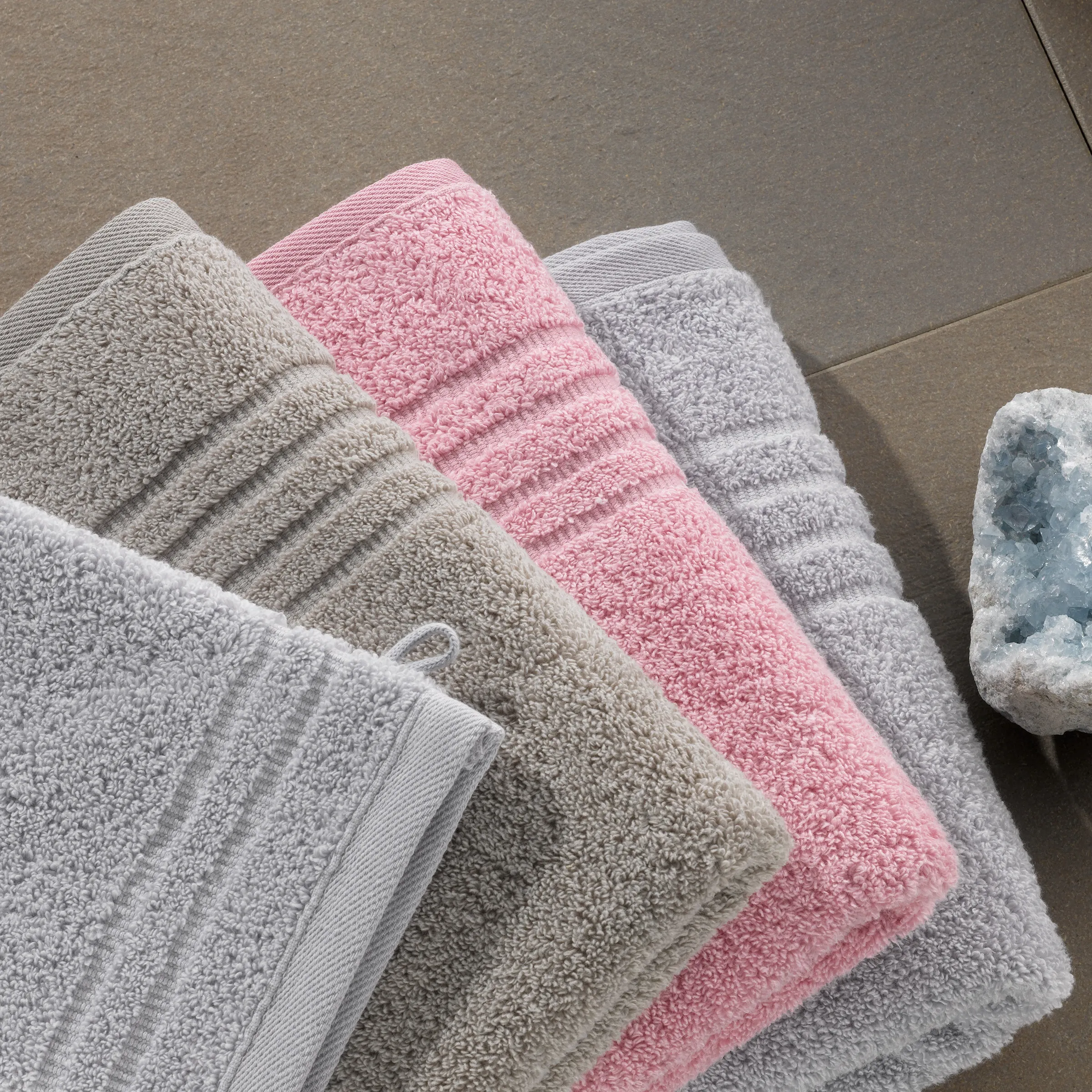 Hochwertige Handtücher Set - Baumwolle in verschiedenen Farben Stapel von weichen Handtüchern in Creme, Beige, Rosa und Grau mit geripptem Rand auf grauem Untergrund